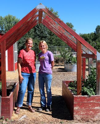 two smiling women, both wearing jeans and short-sleeved shirts, stand under a red wooden frame with raised beds on either side containing green plants