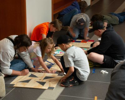 group of children and one woman sit on the floor painting geometric shapes onto squares of plywood