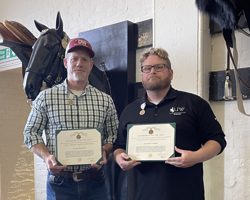 two men, one wearing red University of Wyoming baseball cap and the other wearing a UW Extension shirt, stand together in front of a horse sculpture mounted on a wall. A medal is pinned to the right of each man's shirt collar and they are both holding award plaques from the U.S. Department of the Army.