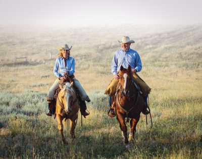 A woman and man on horseback. They are wearing matching blue shirts, cowboy hats, and chaps and are both white. 