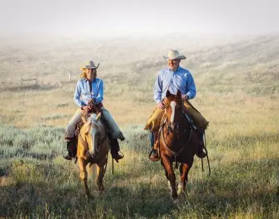 A woman and man on horseback. They are wearing matching blue shirts, cowboy hats, and chaps and are both white.