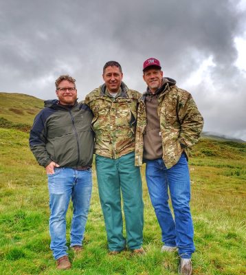 three smiling men, two wearing camouflage jackets, stand together facing the camera with lush green grass and a cloudy sky behind them.