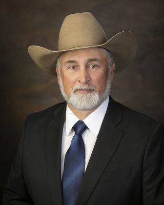 A white man with a short white beard and blue eyes wearing a cowboy hat and black suit. 