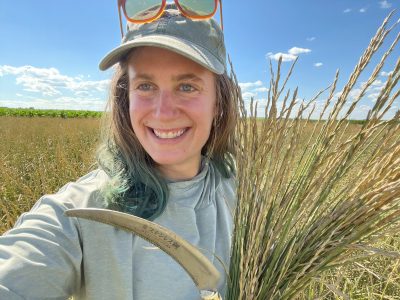 A woman wearing a baseball cap and holding a scythe and a bundle of wheat-like plants.