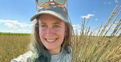 A woman wearing a baseball cap and holding a scythe and a bundle of wheat-like plants.