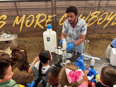 A man with brown skin and black hair demonstrates an experiment to children. He has several large test tubes, one which is full of bubbles, a bowl of soapy water, and a large jug. Kids are wearing rubber gloves, some of which are covered in bubbles.