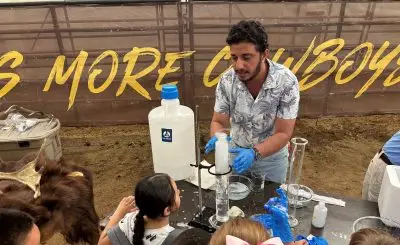 A man with brown skin and black hair demonstrates an experiment to children. He has several large test tubes, one which is full of bubbles, a bowl of soapy water, and a large jug. Kids are wearing rubber gloves, some of which are covered in bubbles.