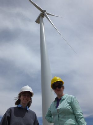 Two women in hard hats underneath a wind turbine. 