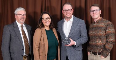 A group of three men and a woman in semi-formal outfits. A young man wearing a gray suit jacket, glasses, and jeans is holding a small circular glass award. 