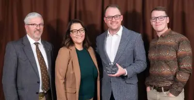 A group of three men and a woman in semi-formal outfits. A young man wearing a gray suit jacket, glasses, and jeans is holding a small circular glass award.