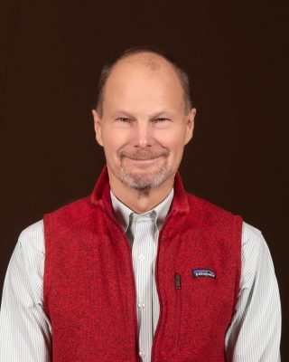 A man with a short gray beard and short gray hair wearing a collared shirt and red vest. 