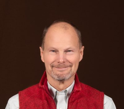 A man with a short gray beard and short gray hair wearing a collared shirt and red vest.