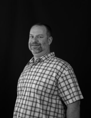 A man with cropped gray hair and a short beard wearing a plaid shirt. The picture is grayscale. 