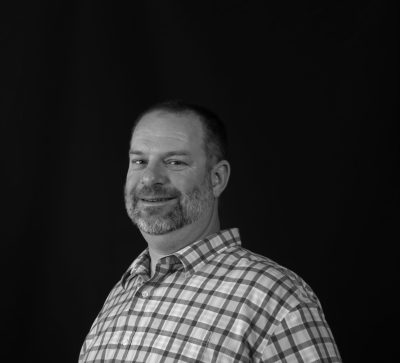 A man with cropped gray hair and a short beard wearing a plaid shirt. The picture is grayscale.