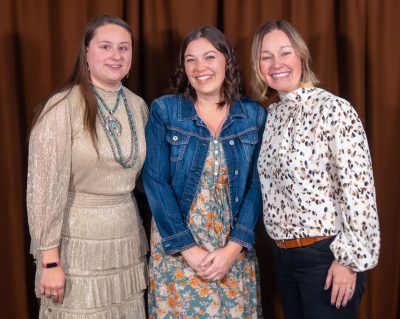 A group of three women in semi-formal outfits. The woman in the middle has bobbed, curled brown hair and is wearing a jean jacket and a long dress with a yellow floral pattern. 