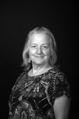 A woman with fine shoulder length straight gray hair wearing a brightly patterned blouse.