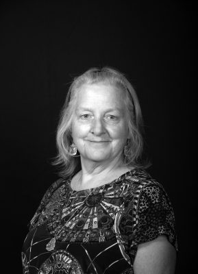 A woman with fine shoulder length straight gray hair wearing a brightly patterned blouse.