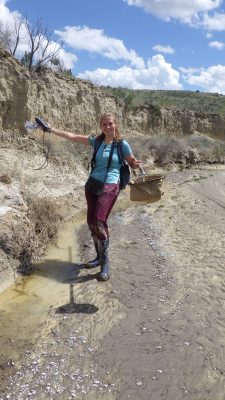 A smiling young woman holding out a net and other equipment. She is wearing rubber books with mud on them, a backpack, and a purse-like thing around her neck. She is walking by very shallow stream of water along the outside of a muddy streambed. 