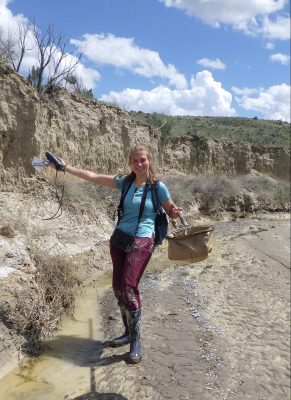 A smiling young woman holding out a net and other equipment. She is wearing rubber books with mud on them, a backpack, and a purse-like thing around her neck. She is walking by very shallow stream of water along the outside of a muddy streambed.