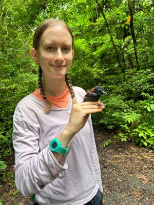 A woman with straight blonde hair braided into two shoulder-length braids. She is holding a small dark brown bird with lighter brown feathers on its back and a blue ring around its eye. The background is lush and green.