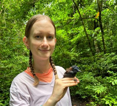 A woman with straight blonde hair braided into two shoulder-length braids. She is holding a small dark brown bird with lighter brown feathers on its back and a blue ring around its eye. The background is lush and green.