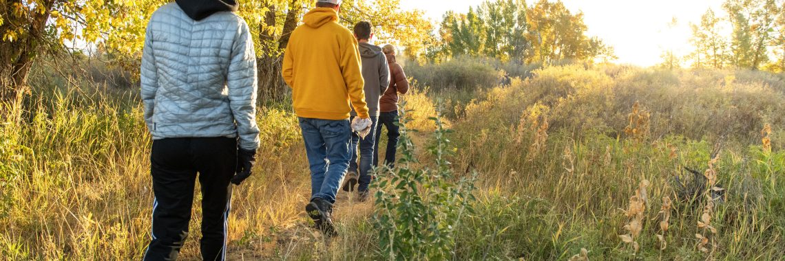 Four people in warm clothing walk down a path in a wooded, grassy area. They are surrounded by fall sunshine and golden fall foliage.