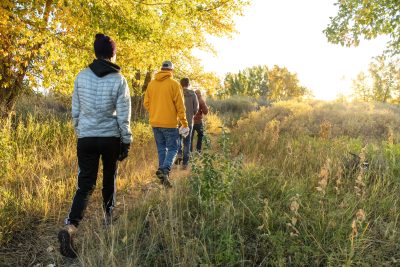 Four people in warm clothing walk down a path in a wooded, grassy area. They are surrounded by fall sunshine and golden fall foliage.