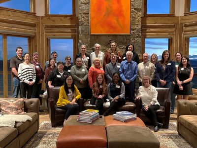 24 people stand in front of a fireplace in a room with several couches and large windows.