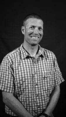 A black and white photo of a man with cropped hair and bear wearing a checked short-sleeved collared shirt. 