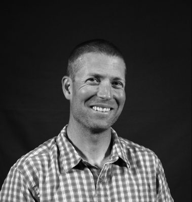 A black and white photo of a smiling man wearing a checked short-sleeved collared shirt.