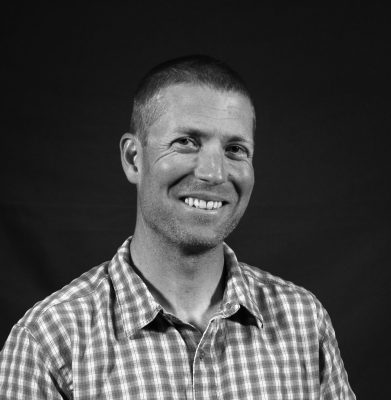 A black and white photo of a smiling man wearing a checked short-sleeved collared shirt.