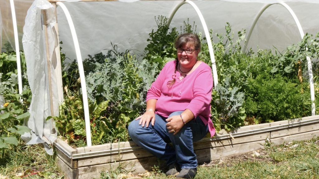 smiling woman wearing glasses and a pink shirt and jeans sits on the wooden edge of a high tunnel full of lush green plants