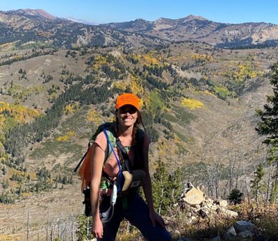 A young woman wearing a ballcap and backpack on a trail in the mountains. 