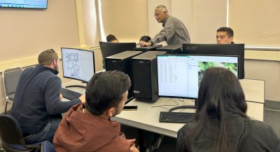 Six young people in a computer lab while an older man leans over one of their screens. 