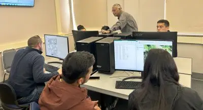 Six young people in a computer lab while an older man leans over one of their screens.