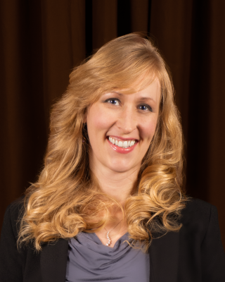 A woman with wavy shoulder length blonde hair wearing a gray shirt and black jacket.