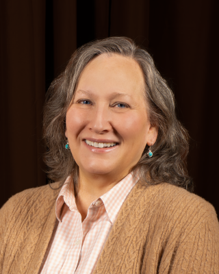 A woman with wavy bobbed gray hair wearing a brown cardigan and a light pink collared shirt. 