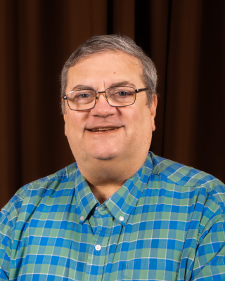 A man with short gray hair and glasses wearing a checked blue and green collared shirt. 