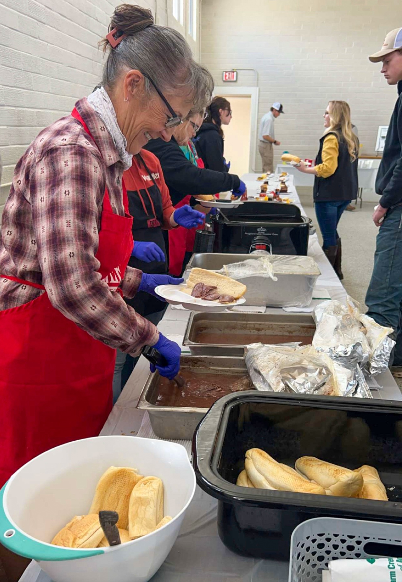 smiling people wearing red aprons and blue food service gloves stand behind a long table and dish food onto plates