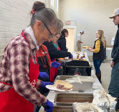 smiling people wearing red aprons and blue food service gloves stand behind a long table and dish food onto plates