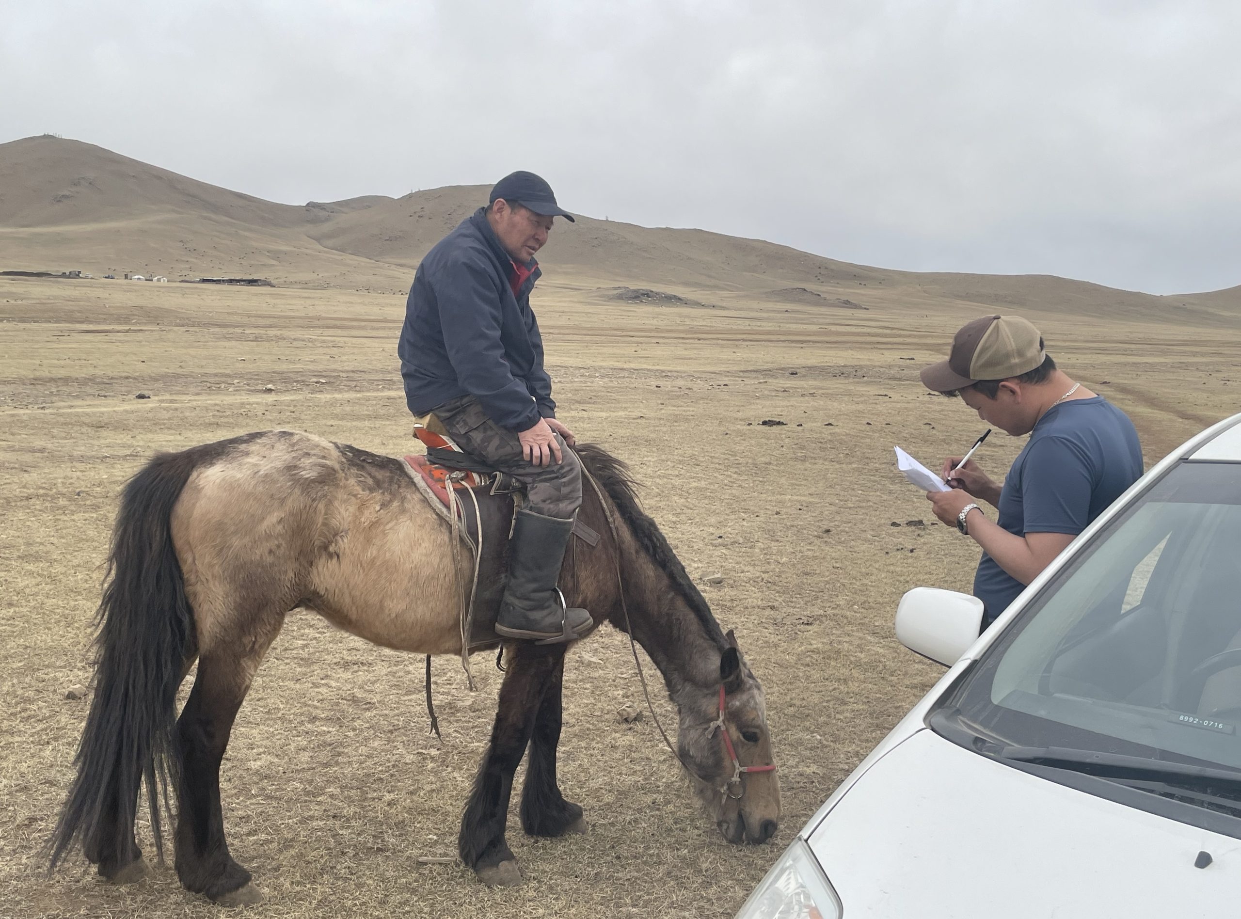 in a barren brown landscape with slopes in the distance, a man on horseback converses with a man leaning against the front of a car while taking notes