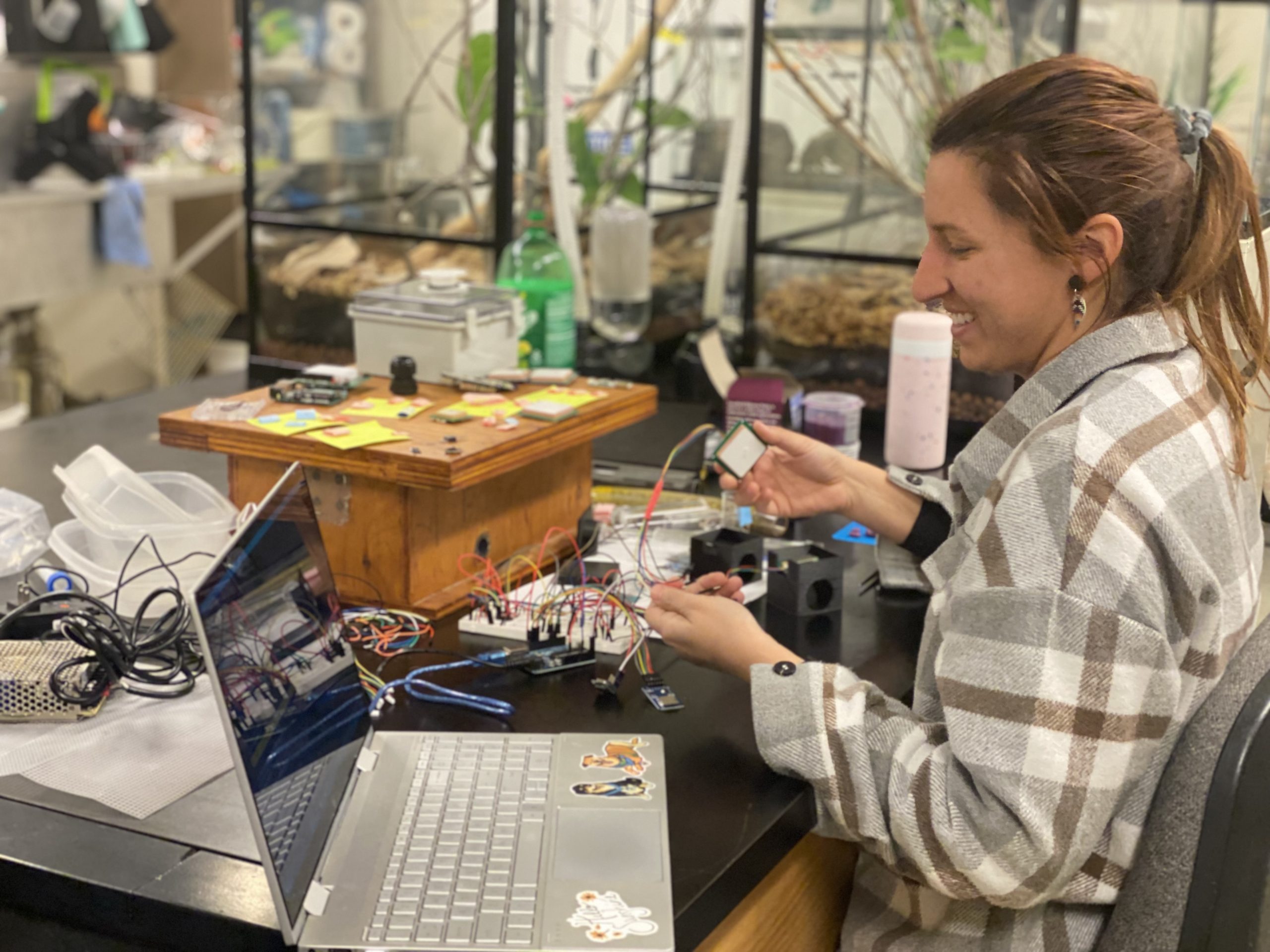 Smiling woman holds small colorful wires connected to tiny electronic devices. An open laptop sits on the table beside her.