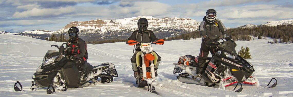 Three helmeted winter recreationists, two riding snowmobiles and one riding a dirt bike with ski tracks, pause on a snow-covered expanse bordered by pines and mountains