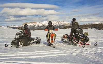 Three helmeted winter recreationists, two riding snowmobiles and one riding a dirt bike with ski tracks, pause on a snow-covered expanse bordered by pines and mountains