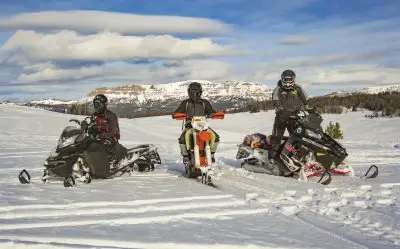 Three helmeted winter recreationists, two riding snowmobiles and one riding a dirt bike with ski tracks, pause on a snow-covered expanse bordered by pines and mountains