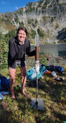 A woman with long brown hair next to a giant plastic vial filled with brown liquid on a metal screw pole. Behind her, various items are strewn on the shore of a mountain lake. 