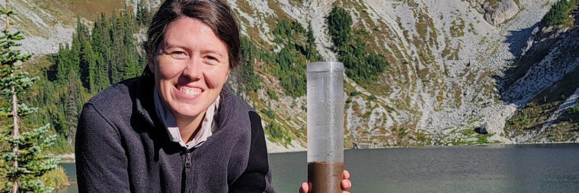 A woman with long brown hair next to a giant plastic vial filled with brown liquid on a metal screw pole. Behind her, various items are strewn on the shore of a mountain lake.