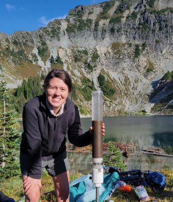 A woman with long brown hair next to a giant plastic vial filled with brown liquid on a metal screw pole. Behind her, various items are strewn on the shore of a mountain lake.