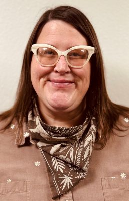 A woman with straight brown shoulder-length hair wearing a brown scarf with white leaf patterns and chunky cat-eye glasses.