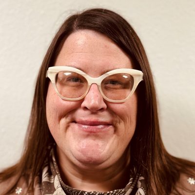 A woman with straight brown shoulder-length hair wearing a brown scarf with white leaf patterns and chunky cat-eye glasses.